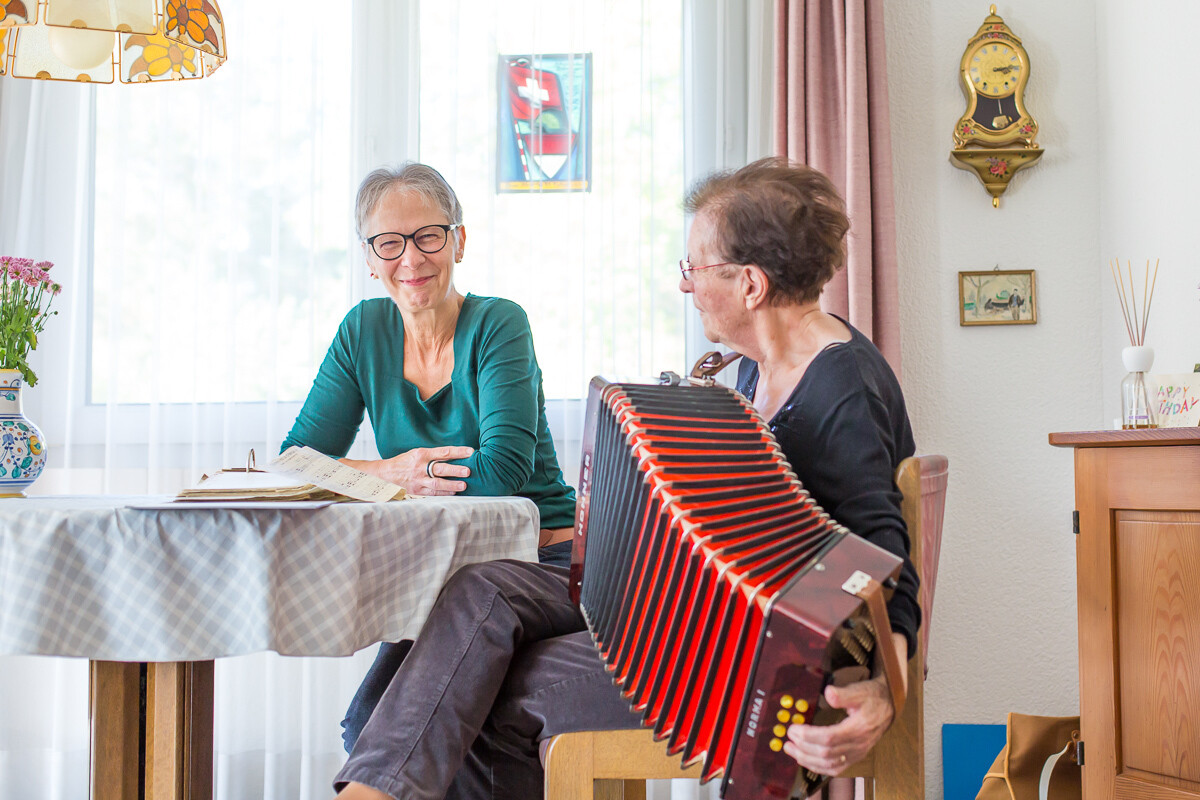 Eine ältere Frau spielt an einem Tisch Akkordeon, während ihre lächelnde Betreuerin ihr zuhört.
