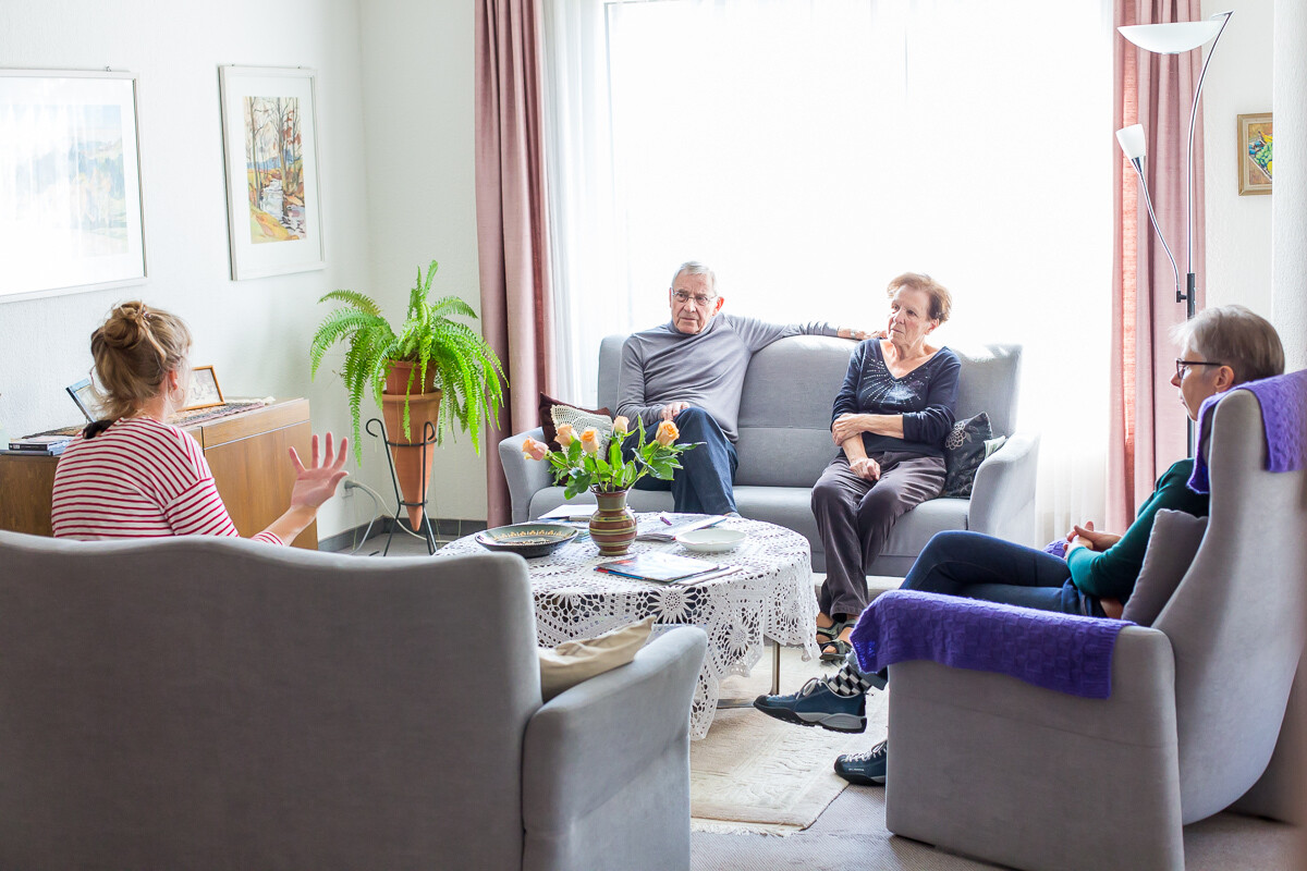 Ein älteres Ehepaar sitzt in einem hellen Wohnzimmer auf dem Sofa und sprechen mit zwei Frauen vom Entlastungsdienst